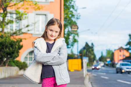 Outdoor portrait of a cute little girl in a city wearing warm grey knitted jacket standing next to the roadの写真素材