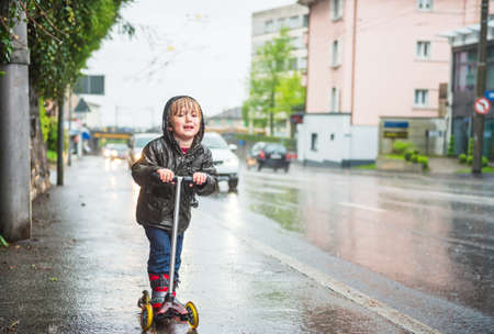 Cute little boy in a very wet clothes on the scooter next to road under the rainの写真素材