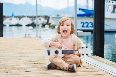 Little happy boy plays his guitar or ukulele, sitting by the lake