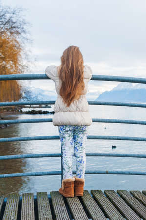 Cute little girl standing on a bridge, looking at the beautiful lake, wearing warm beige coat, back viewの写真素材