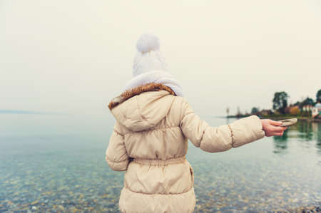 Little girl playing by the lake, throwing stones into the water on a nice cold day, wearing warm white hat, scarf and beige coat, back view, toned imageの写真素材