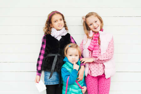 3 cute little girls against white wooden background, wearing warm waistcoatsの写真素材