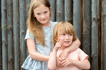 Outdoor portrait of two cute kids, big sister and her little brother against wooden wallの写真素材