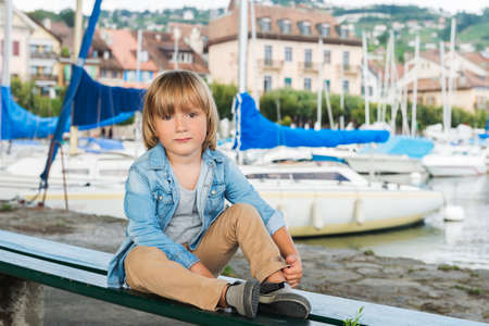 Fashion portrait of a cute little blond boy sitting on a bench by the lake, wearing denim shirt and beige trousersの写真素材