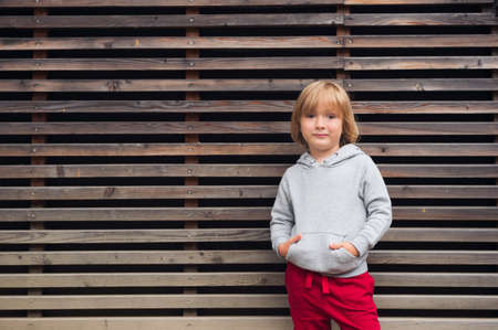 Fashion portrait of adorable toddler boy wearing grey sweatshirt and red trainings, standing against...