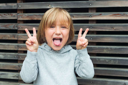 Close up portrait of a funny little boy, wearing grey sweatshirt, pulling a tongue and gesturing peace signの写真素材