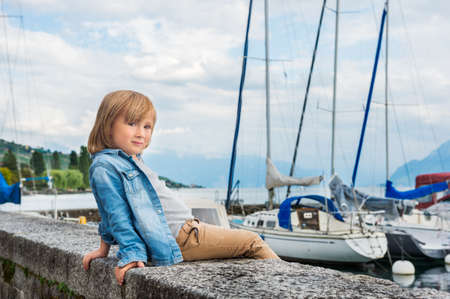 Fashion portrait of a cute little blond boy sitting by the lake, wearing denim shirt and beige trousersの写真素材