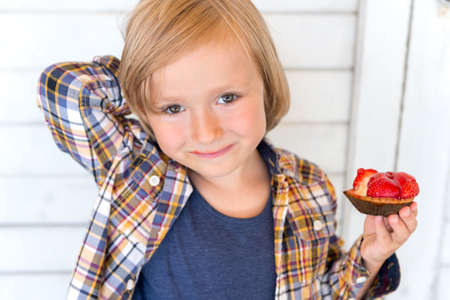 Close up portrait of adorable kid boy, standing against white wooden background, holding strawberry cakeの写真素材