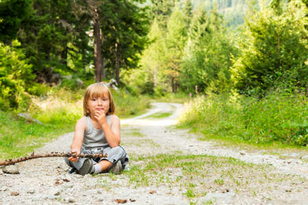 Portrait of a cute little boy hiking in a forest, sitting on ground, holding a stickの写真素材
