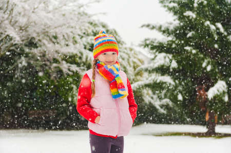 Winter portrait of a cute little girl under the snowfall, wearing red pullover, colorful hat and scarfの写真素材