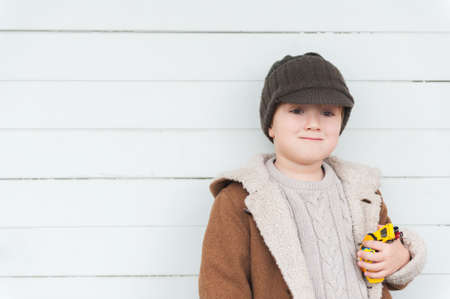 Portrait of a cute little boy against white wooden background, wearing warm beige pullover, coat and brown hat, holding toy carsの写真素材