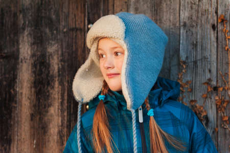 Close up portrait of a cute little girl against wooden background, wearing winter hat and warm blue...