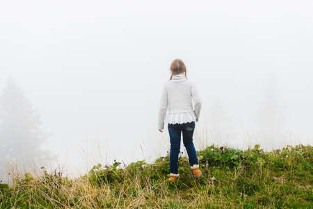 Cute little girl of 8 years old playing outdoors on a very foggy day, wearing grey warm pullover,...