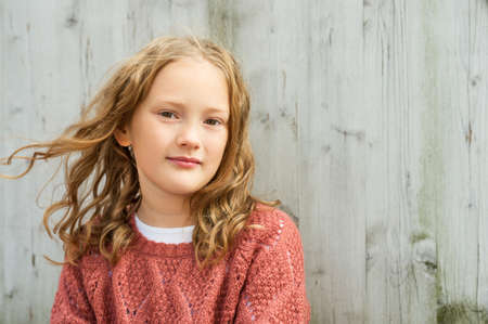 Close up portrait of a cute little girl of 8 years old with curly hair, wearing terracotta pullover, sitting against grey wooden backgroundの写真素材