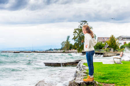 Cute little girl of 8 years old playing by the lake on a very windy day, wearing warm white knitted pulloverの写真素材
