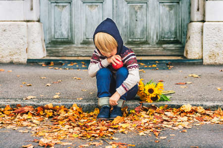 Autumn portrait of adorable little blond boy of 4 years old, wearing warm pullover with the hood, dark denim jeans and blue shoes, holding an apple and bouquet of yellow sunflowersの写真素材