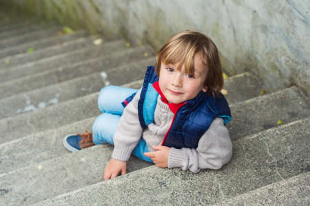 Fashion portrait of a cute little blond boy sitting on stairs in a city, wearing beige pullover and blue jacketの写真素材
