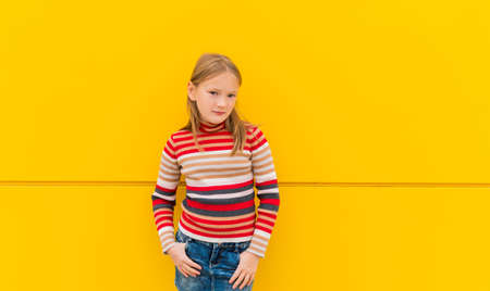 Portrait of a cute little girl of 8 years old, wearing stripe roll neck pullover and denim skirt, standing next to bright yellow wallの写真素材