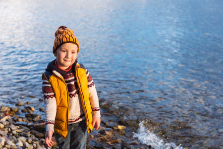 Cute little boy playing next to lake on a cold day, wearing warm pullover, yellow vest and hatの写真素材