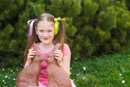 Outdoor portrait of a cute little girl playing in a garden with two chocolate bunniesの写真素材