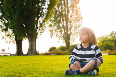Portrait of beautiful smiling cute little boy. 3-4 years old little child playing outside in green summer or spring park. Boy sitting alone on green grass at sunset, wearing stripe jersey sweatshirtの写真素材