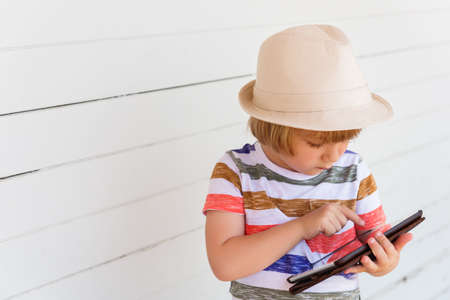 Cute little boy playing with tablet pc wearing a hat, standing against white wooden backgroundの写真素材