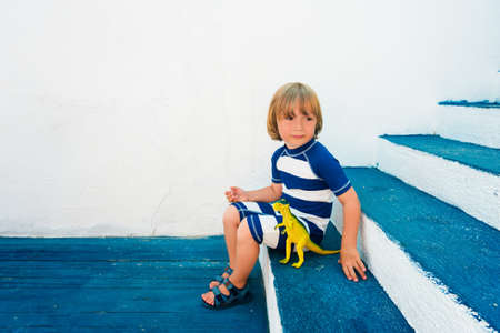 Adorable little boy playing with a dinosaur toy, wearing funny swim suit and beach shoes, sitting on blue stairs, summer vacations conceptの写真素材
