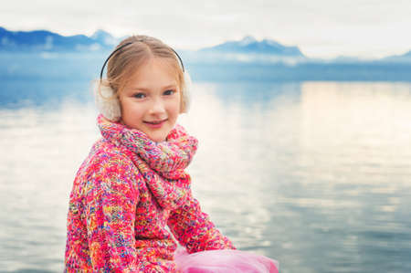 Outdoor close up portrait of adorable little kid girl of 8 years old resting by the lake Geneva on a nice winter day, wearing colorful pink pullover and warm earmuffsの写真素材