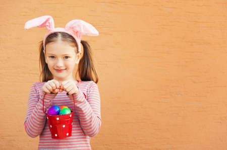 Cute little girl wearing rabbit ears, holding bucket with colorful easter eggsの写真素材