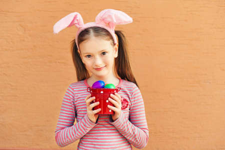 Cute little girl wearing rabbit ears, holding bucket with colorful easter eggs