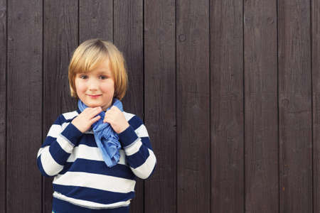 Fashion portrait of adorable kid boy against dark brown wooden background, wearing white and blue...