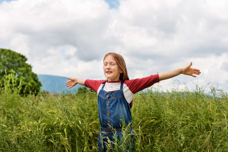 Little girl playing in green wheat field in summertime, wearing red and white t-shirt and overalls, arms wide openの写真素材