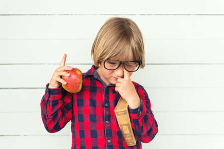 Tired little schoolboy holding book and red apple, wearing white red and blue plaid shirt and black frame eyeglasses, standing against white wooden background. Back to school conceptの写真素材