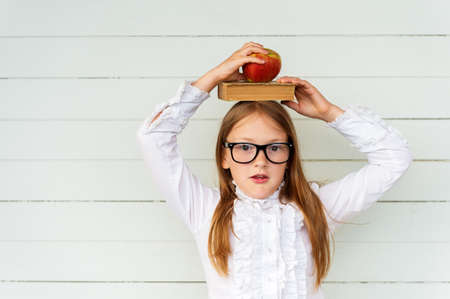 Happy little schoolgirl with book and red apple on her head, wearing white vintage blouse and black frame eyeglasses, standing against white wooden background. Back to school conceptの写真素材