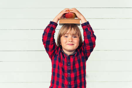 Happy little schoolboy with book and red apple on his head, wearing red and blue plaid shirt, standing against white wooden background. Back to school conceptの写真素材