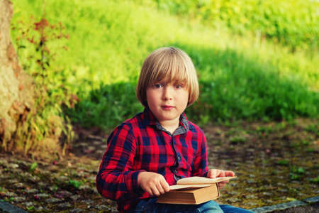 Sweet little boy reading old book in park, wearing red and blue plaid shirt. Toned imageの写真素材