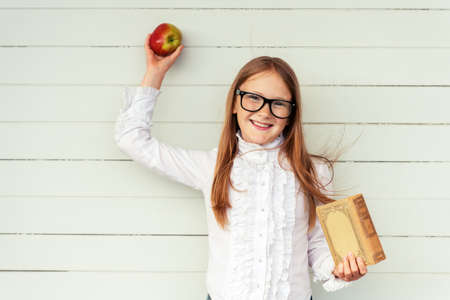 Happy little schoolgirl holding book and red apple, wearing white vintage blouse and black frame eyeglasses, standing against white wooden background. Back to school conceptの写真素材