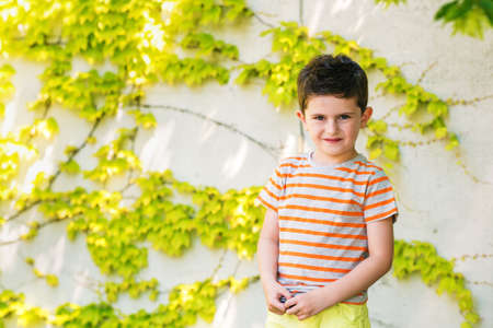 Outdoor portrait of a cute little boy standing against ivy wall on a very sunny day, wearing grey and orange stripe t-shirtの写真素材