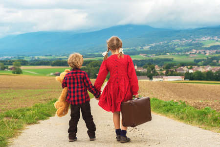 Two cute kids, little girl and her brother, walking down the small road, holding big teddy bear and old brown suitcaseの写真素材