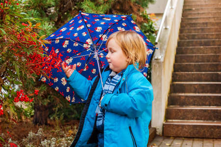 Cute toddler boy with umbrella playing outdoors on a rainy day, wearing blue jacket.の写真素材