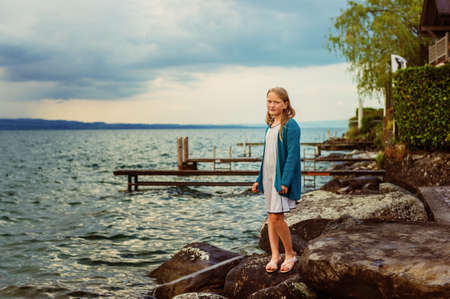 Adorable little girl of 8-9 years old playing by the lake, wearing sandals, dress and blue knitted jacketの写真素材
