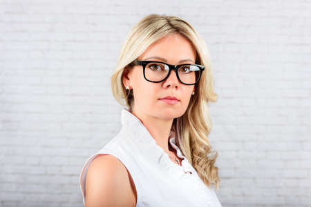 Studio portrait of a young blond woman wearing eyeglasses, standing against white brick background