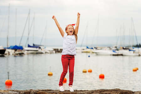Happy girl  9 year old having fun outdoors, playing by lake on a nice warm sunny evening, wearing white t-shirt and shoes, red polka dot trousers and headband. Image taken at Lake Geneva, Switzerlandの写真素材