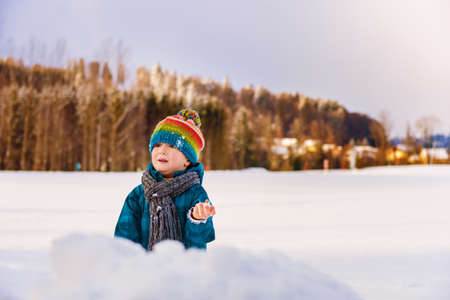 Adorable little boy having fun outdoors on a very cold day, playing snow balls, wearing warm blue waterproof jacket, colorful hat and scarfの写真素材