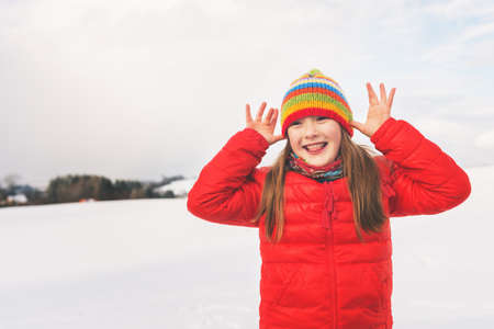 Little girl wearing red jacket and colorful hat, playing with snow in winter timeの写真素材