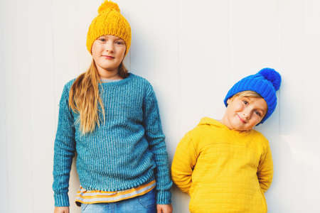 Outdoor portrait of adorable fashion kids, wearing warm pullovers and hats, standing against white wooden backgroundの写真素材