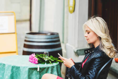 Outdoor portrait of beautiful blond woman holding bouquet of pink roses, resting in cafe, using...