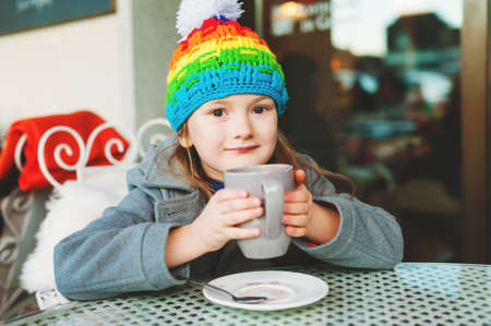 Adorable little 6 year old girl drinking hot chocolate in winter cafe, holding big cup, wearing grey coat and colorful hatの写真素材