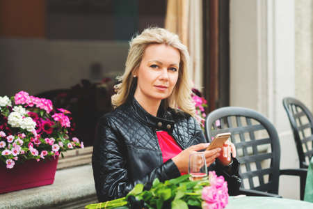 Outdoor portrait of beautiful blond woman holding bouquet of pink roses, resting in cafe, using...