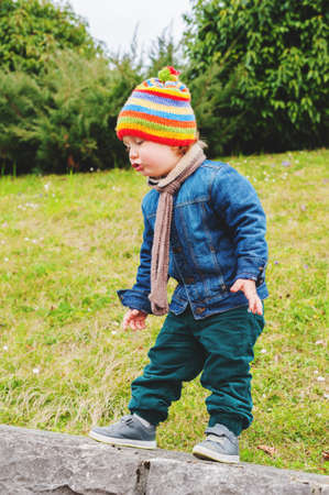Adorable toddler boy playing in park, wearing colorful hat, denim jacket and green trousers